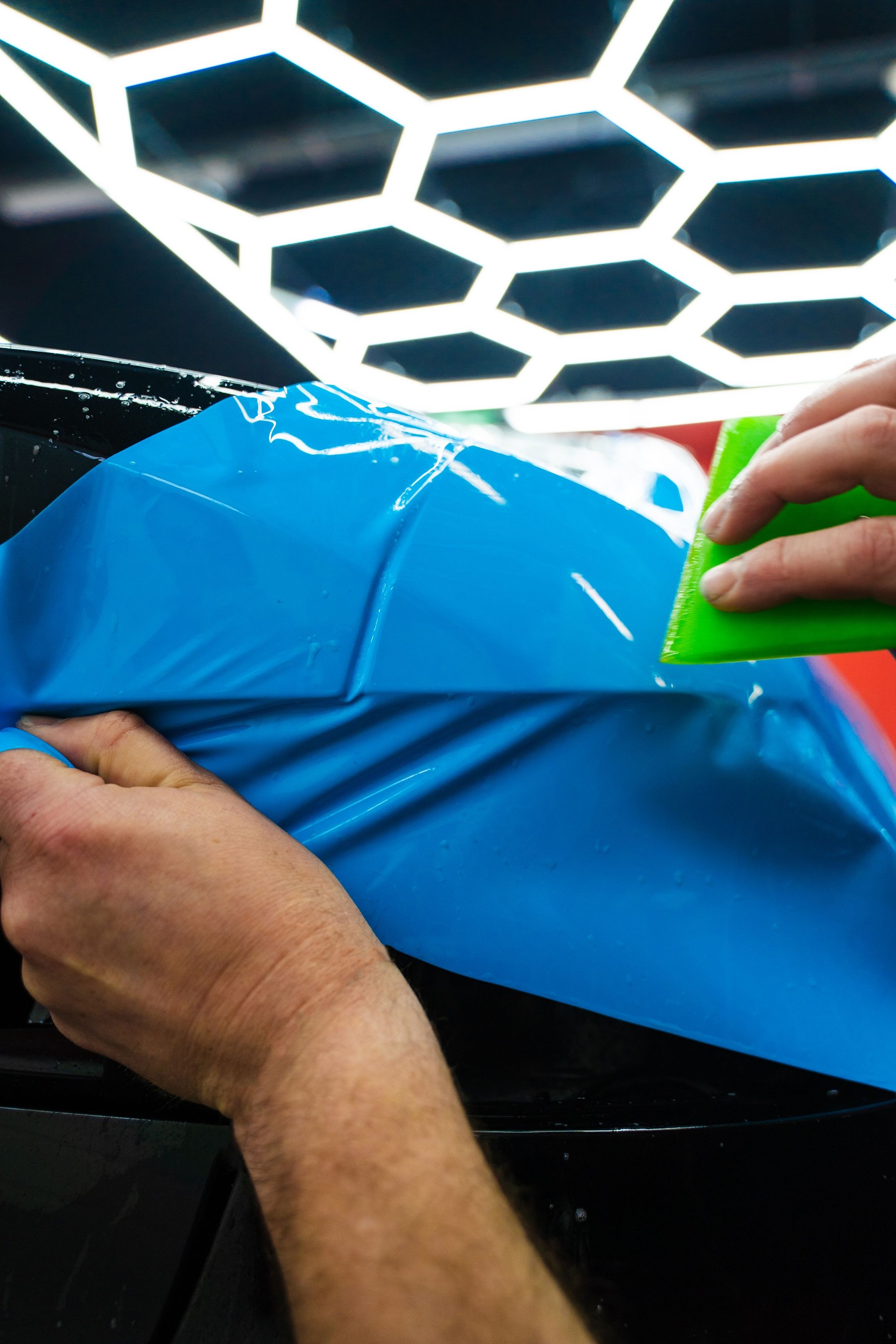 Applying a high-gloss blue PPF using a green squeegee ensures a bubble-free, smooth surface of car. Blue PPF applied with green squeegee during detailing car to preserve finish and block damage.